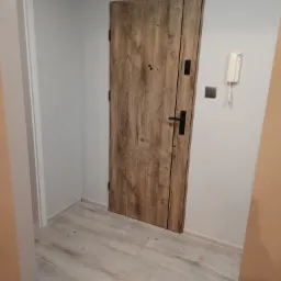 Wejściowe drzwi z drewnopodobnym wzorem, czarna klamka, szara ściana, panele podłogowe imitujące drewno, biały domofon i skrzynka elektryczna w przedpokoju.