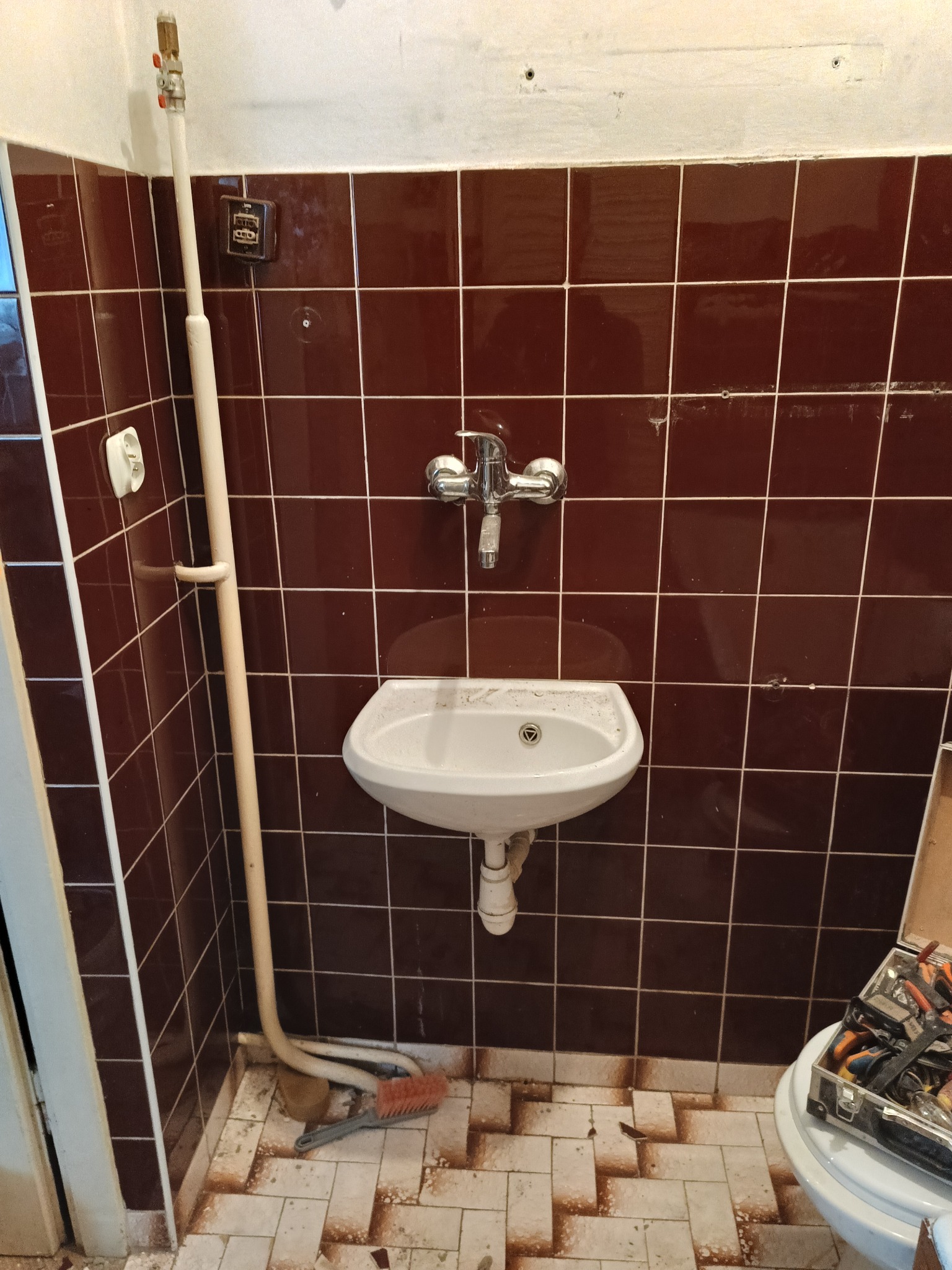 Łazienka w trakcie remontu: brązowe płytki na ścianach, biała umywalka i widoczne narzędzia w kuferku na sedesie. Nieukończone prace remontowe.