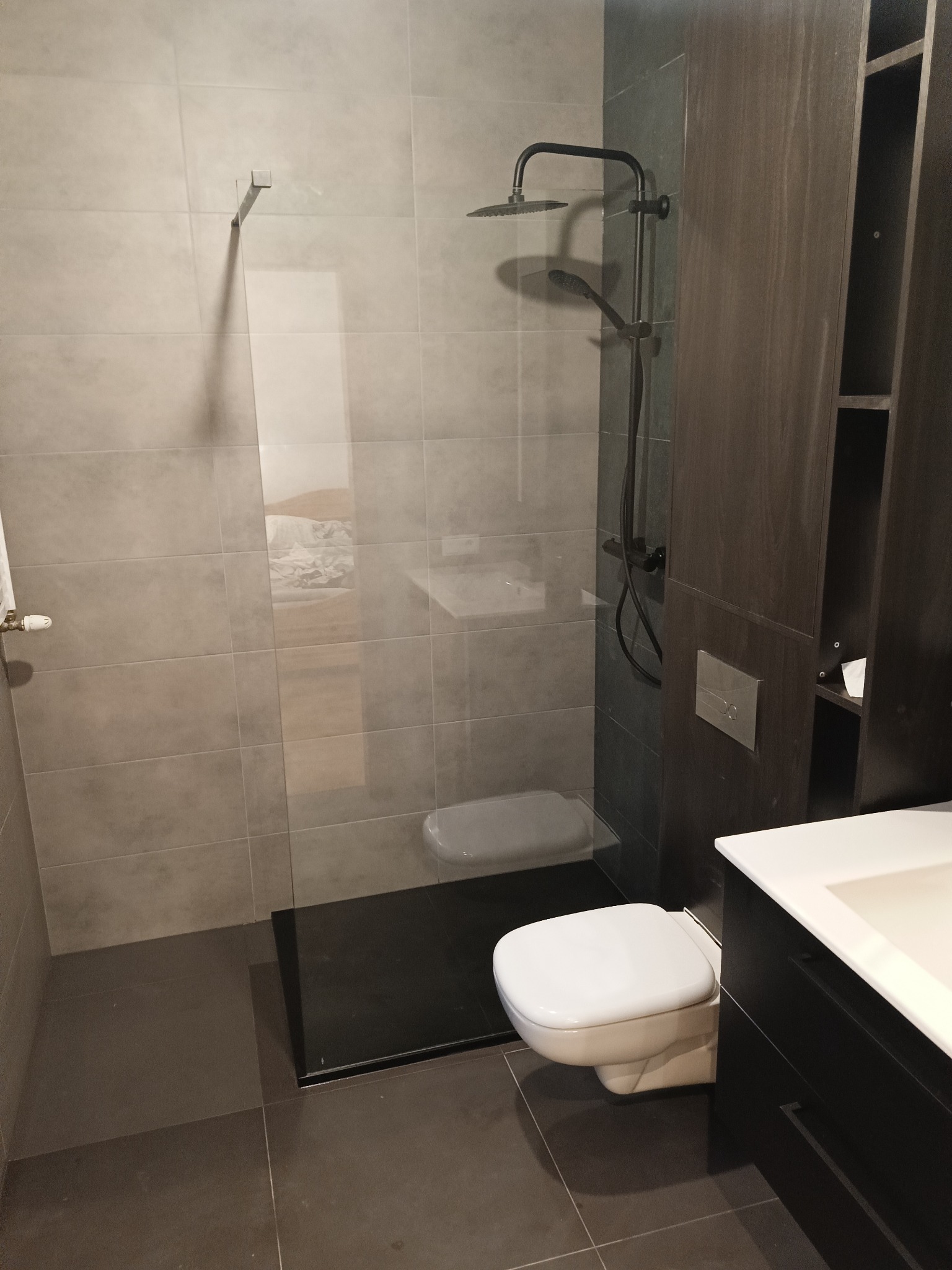 Nowoczesna łazienka z kabiną typu walk-in, szarą glazurą, czarnym brodzikiem i wiszącą toaletą. Minimalistyczny design, ciemne drewno szafki pod umywalką.