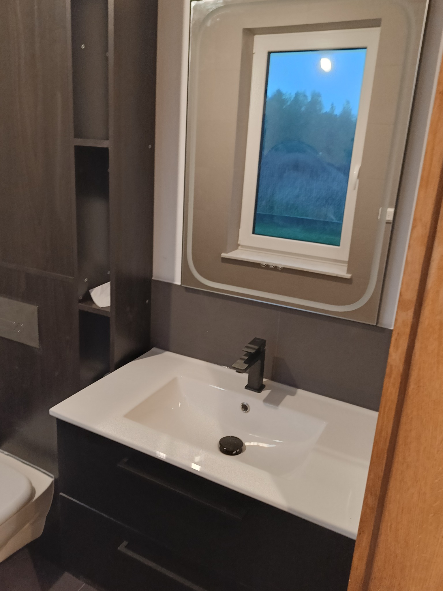 Nowoczesna łazienka: biała umywalka z czarną armaturą i szafką w kolorze grafitu. Nad umywalką lustro z oknem, przez które widać nocny krajobraz.