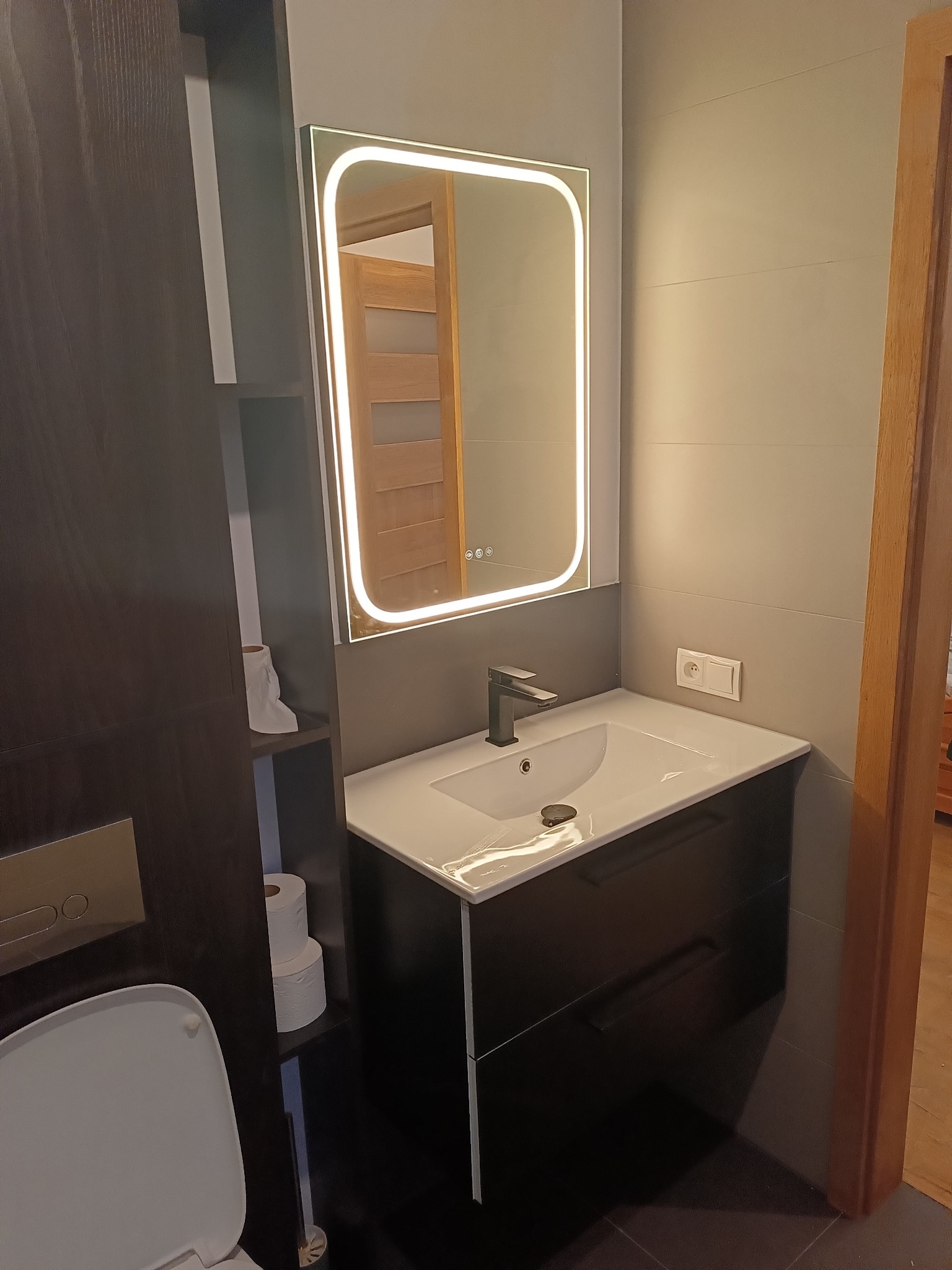 Nowoczesna łazienka z podświetlanym lustrem i umywalką na szafce w odcieniach szarości i czerni. Widoczna toaleta i fragment drzwi.