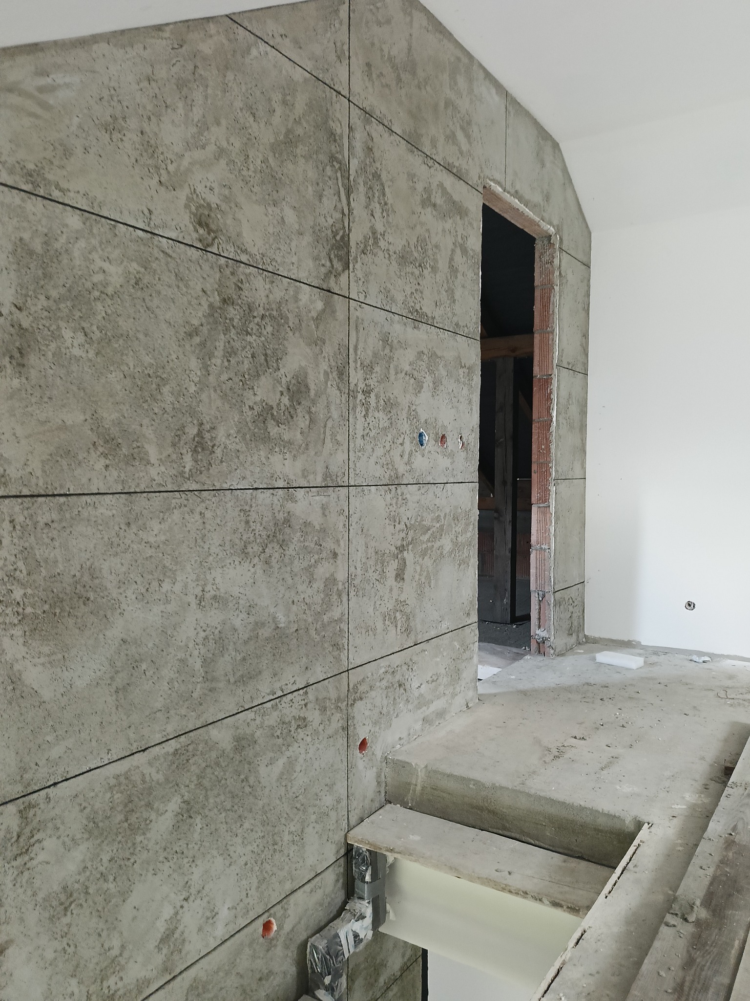 Ściana wyłożona szarymi płytami imitującymi beton z czarnymi fugami. Widoczne otwory na gniazdka i wystający fragment instalacji. W tle surowy otwór drzwiowy.