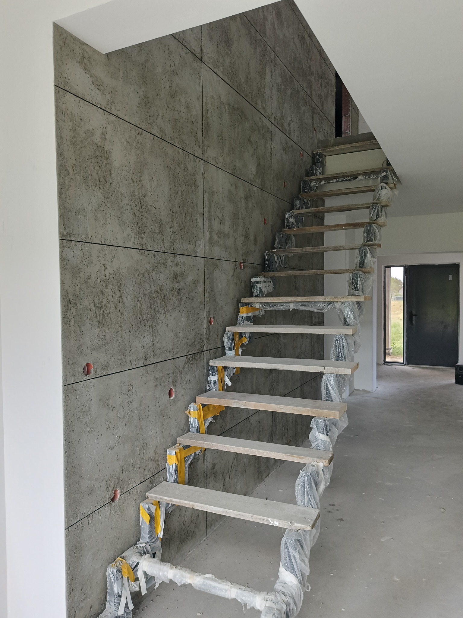 Montaż schodów z drewnianymi stopniami i okładziną ścienną imitującą beton. Schody zabezpieczone folią, widoczne otwory w ścianie. Surowy stan wykończenia wnętrza.