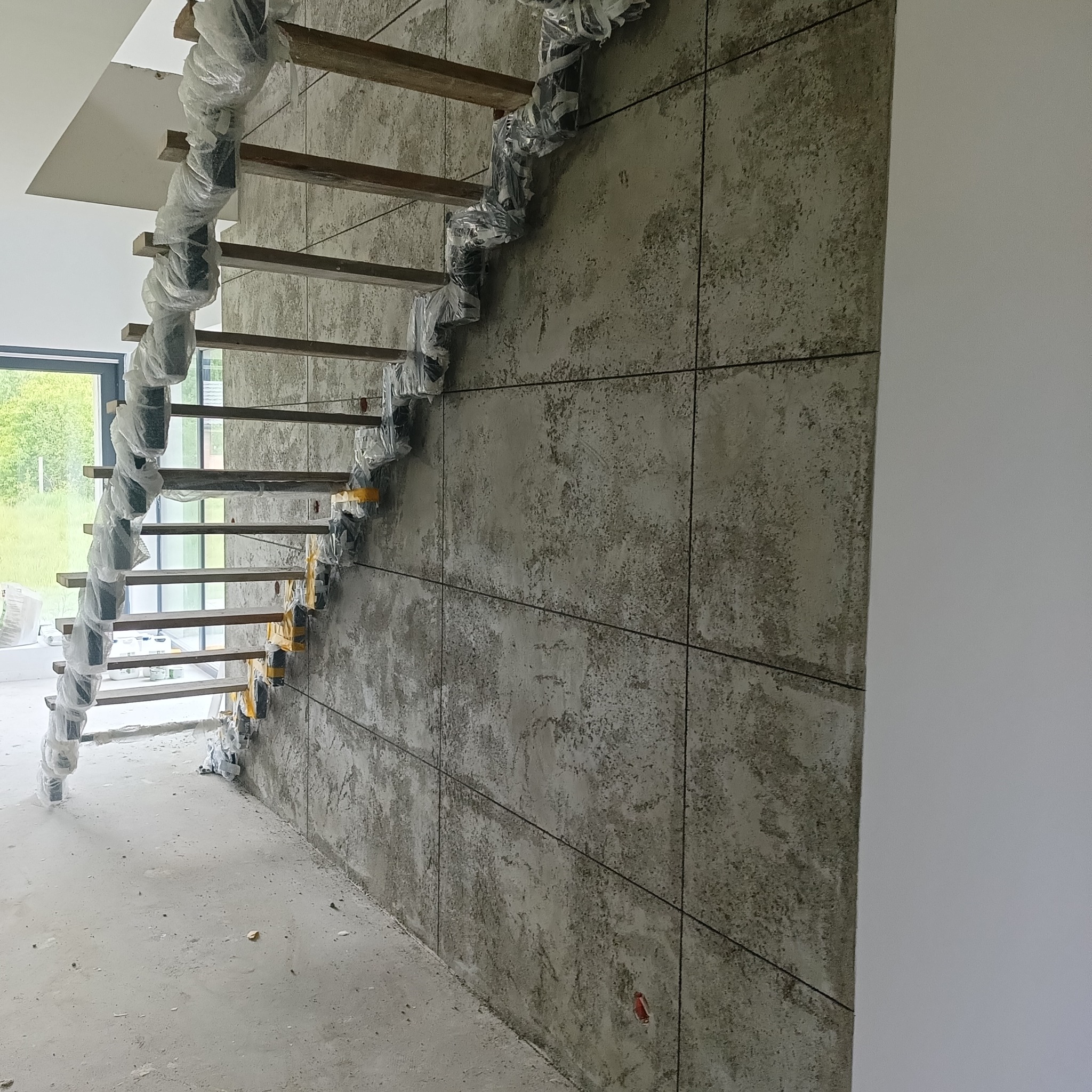 Wnętrze w trakcie wykończenia: spiralne schody z drewnianymi stopniami, zabezpieczone folią, na tle ściany wyłożonej płytami imitującymi beton.