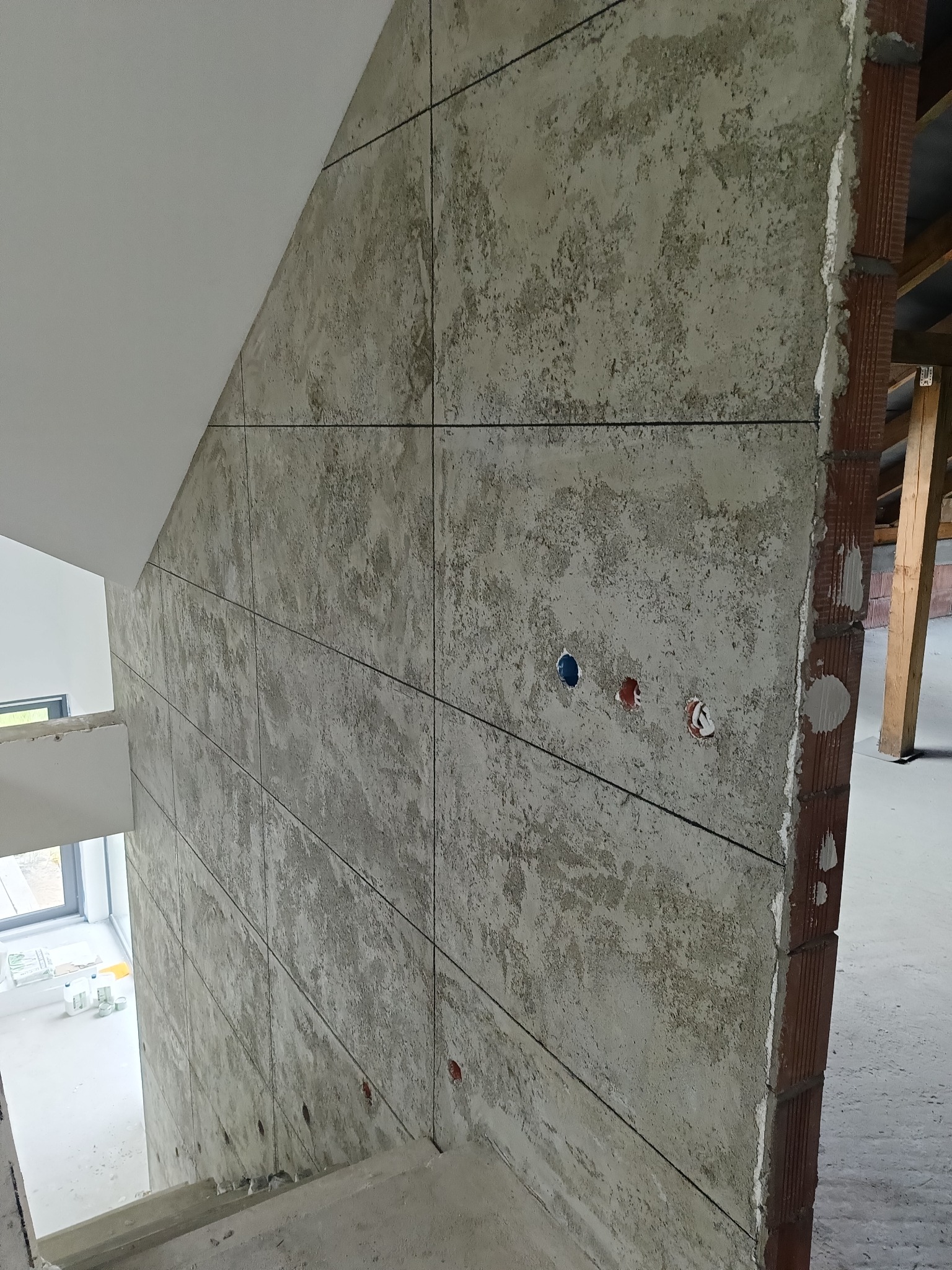 Ściana wykończona płytami imitującymi beton, z widocznymi puszkami elektrycznymi. Surowy, industrialny styl wykończenia wnętrza w trakcie realizacji. Widoczne schody i ceglany fragment ściany.