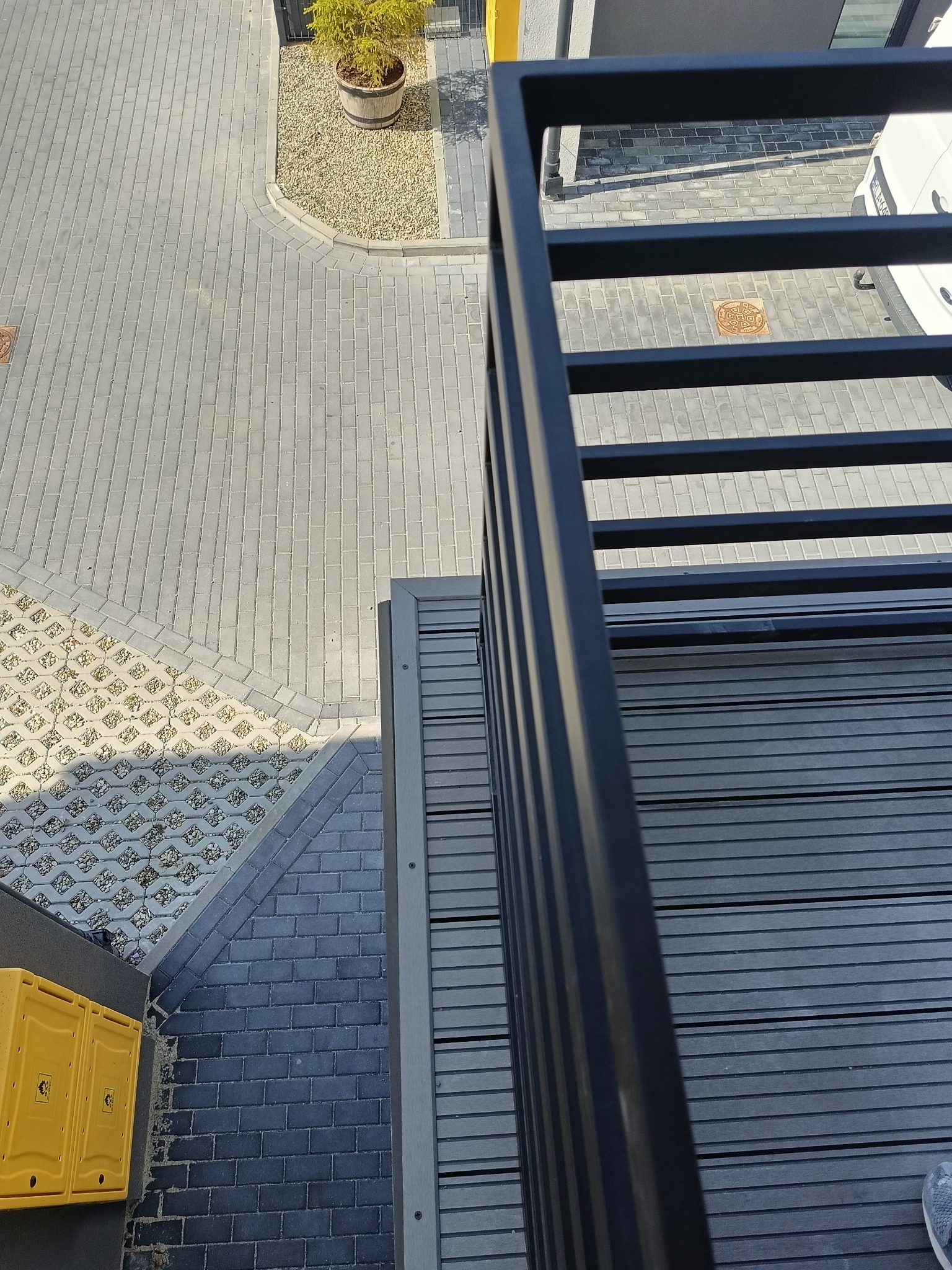 Nowoczesny balkon z szarą, kompozytową podłogą i czarną, metalową balustradą. Widok z góry na podwórze z kostki brukowej i donicę z rośliną.
