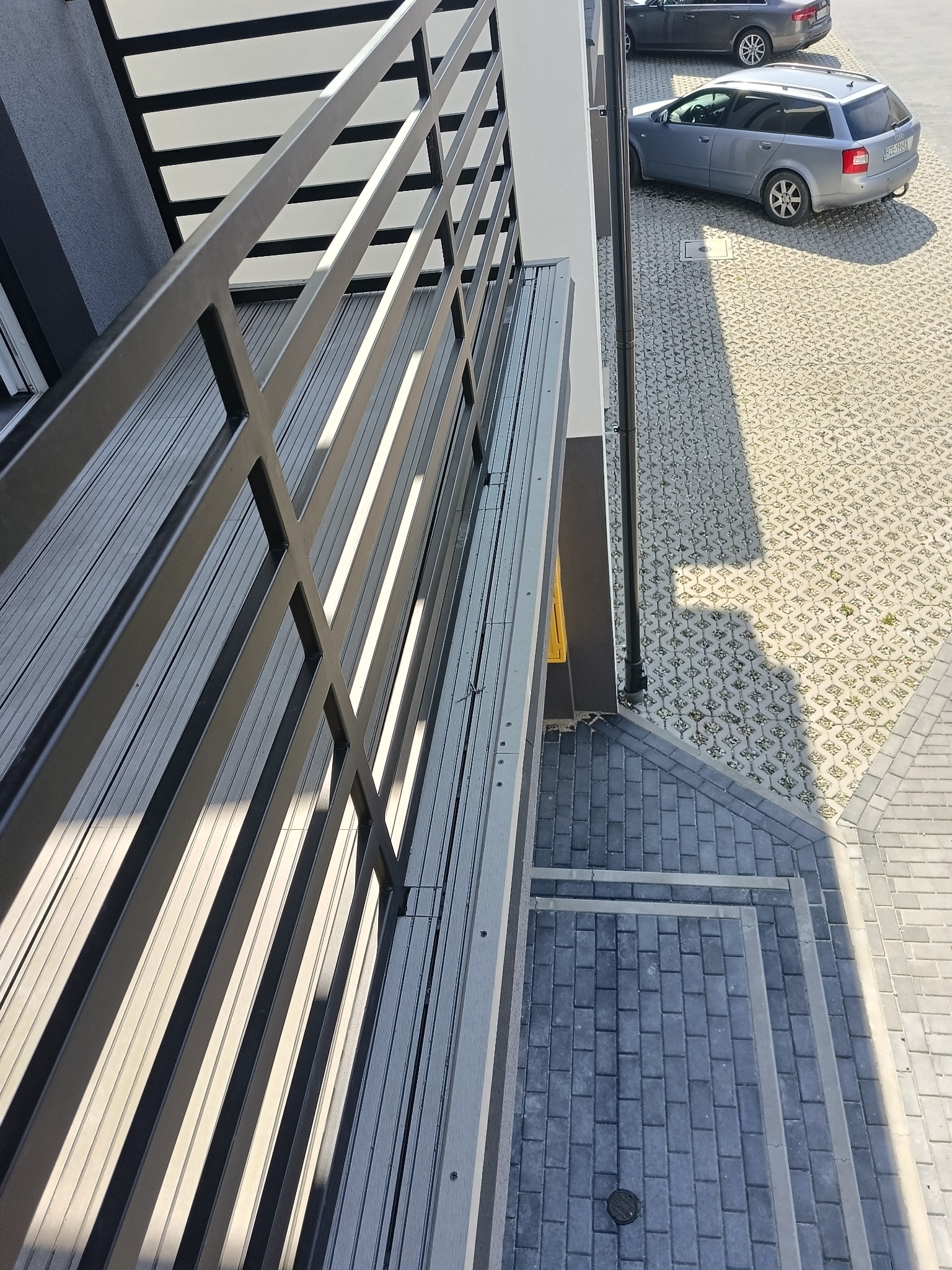 Nowoczesna zabudowa balkonu z szarymi panelami i metalową balustradą, widok z góry na kostkę brukową i zaparkowane auta w tle.