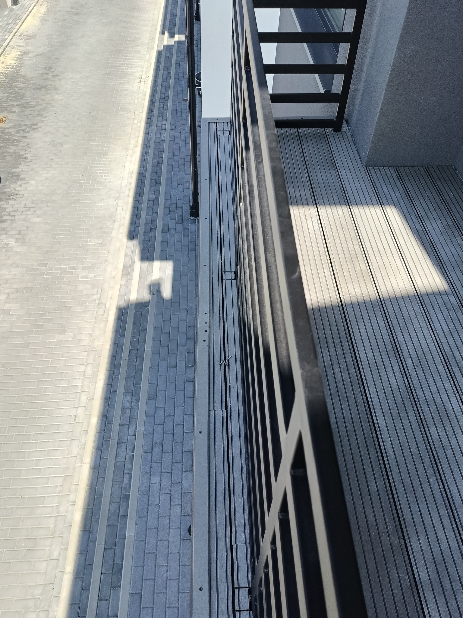 Nowoczesny balkon z szarą deską kompozytową i czarną balustradą, widok z góry, oświetlony słońcem. Elementy schodów na balkonie.