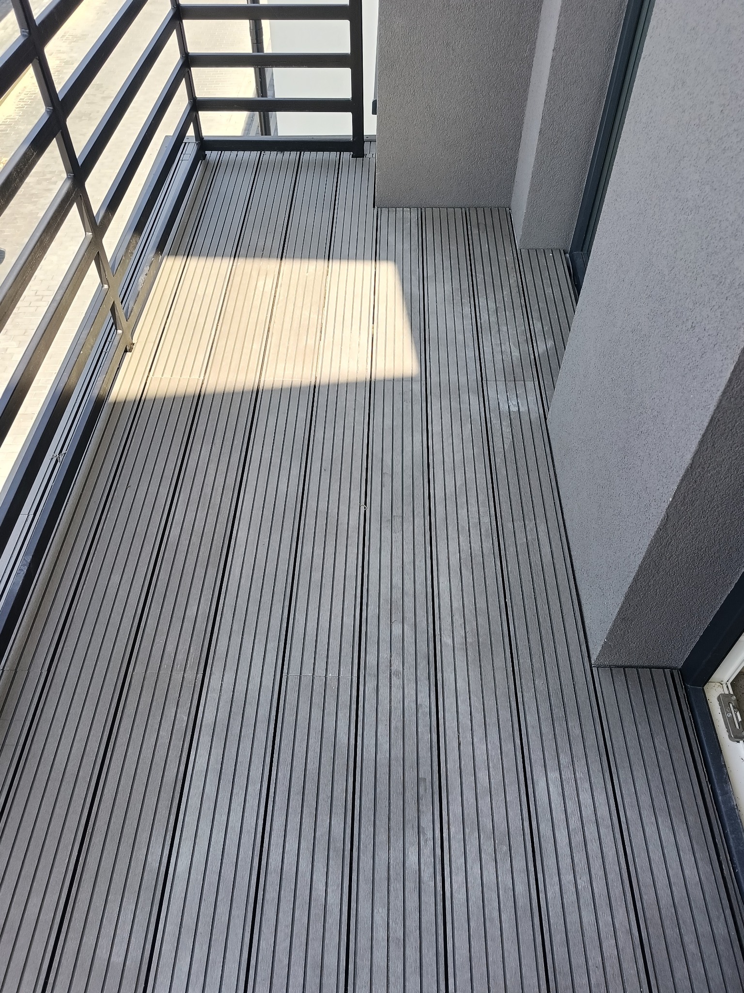 Szare deski kompozytowe na balkonie z czarną balustradą, fragment ściany. Widok z góry, oświetlone naturalnym światłem.