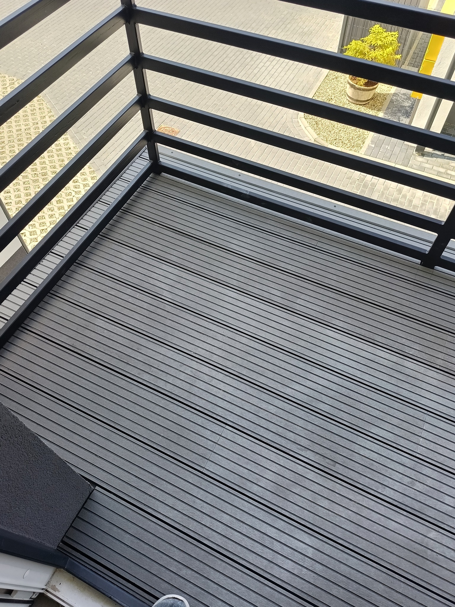 Szare deski kompozytowe na balkonie, widok z góry. Czarne, metalowe barierki i fragment widoku na zewnątrz z rośliną w donicy. Nowoczesna aranżacja balkonu.