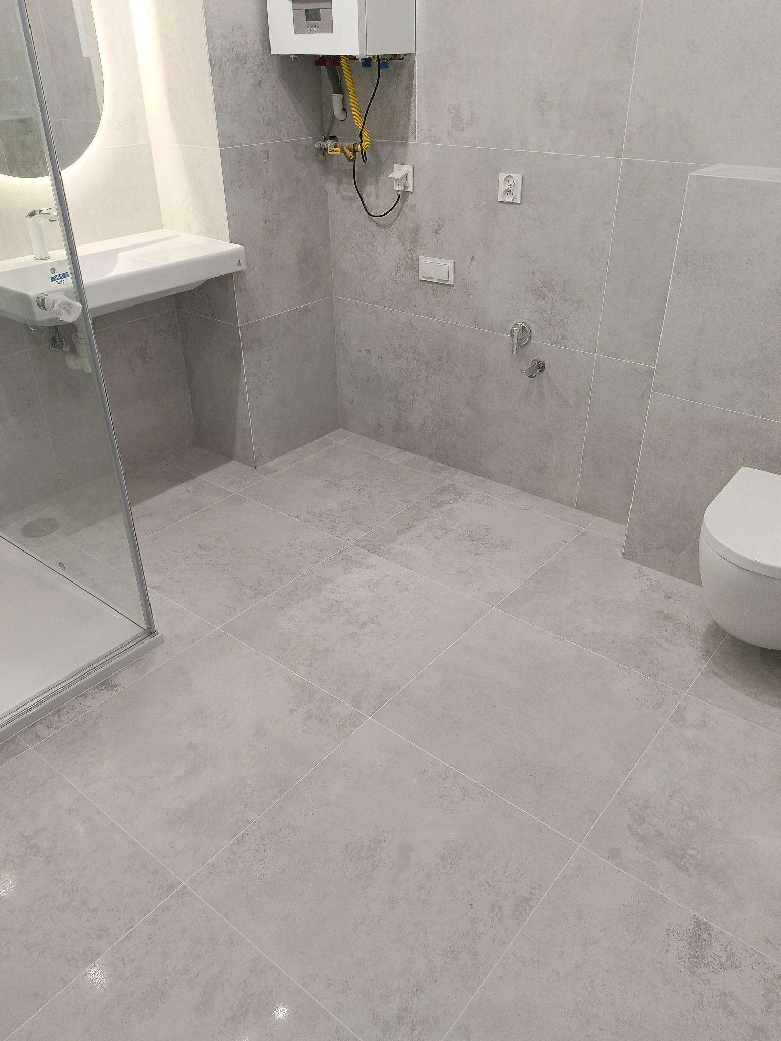 Jasnoszara łazienka po remoncie: kabina prysznicowa, umywalka z okrągłym lustrem, toaleta. Na ścianie piecyk gazowy i gniazdka. Płytki podłogowe i ścienne w jednolitym odcieniu.