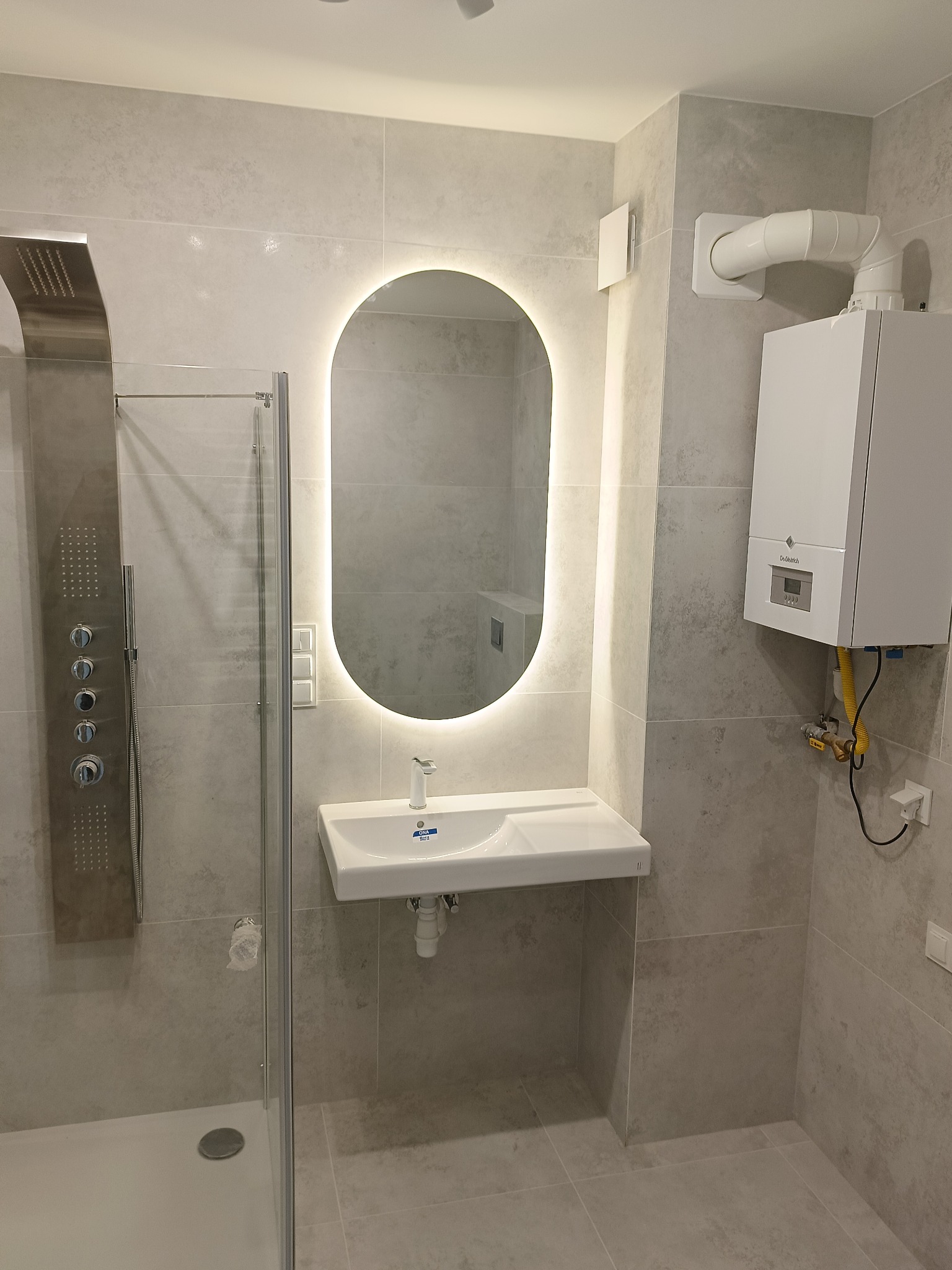 Nowoczesna łazienka w szarościach: kabina prysznicowa z panelem, umywalka z podświetlanym lustrem i piec gazowy na ścianie. Minimalistyczny design.