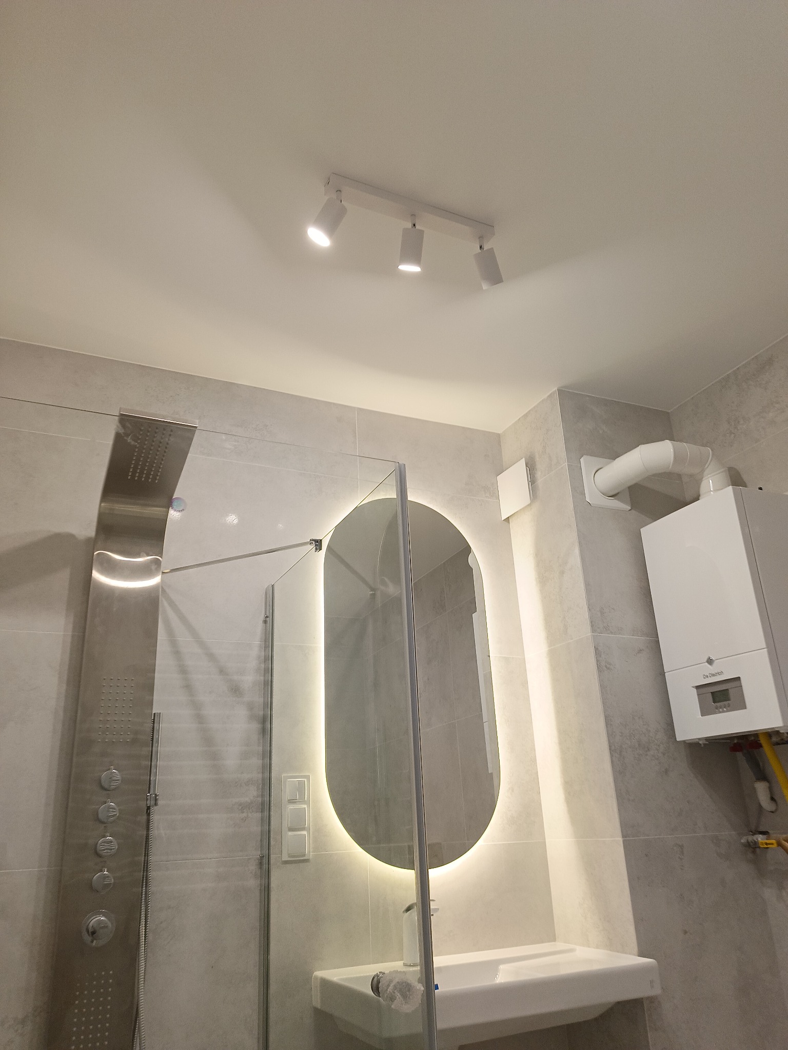 Nowoczesna łazienka z szarymi płytkami, podświetlanym lustrem owalnym i panelem prysznicowym. Widoczny piec gazowy i oświetlenie sufitowe.