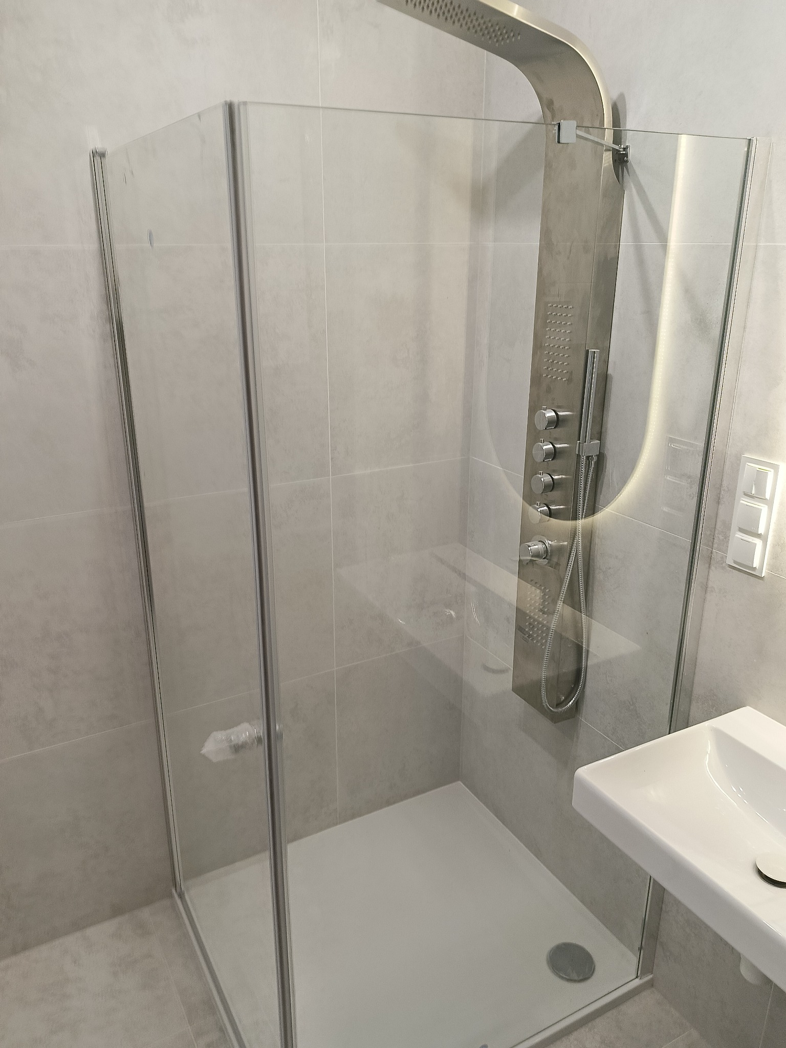 Nowoczesna kabina prysznicowa z deszczownicą i panelem natryskowym, wykończona w szarościach, z widoczną umywalką. Minimalistyczny design, szklane ściany kabiny.