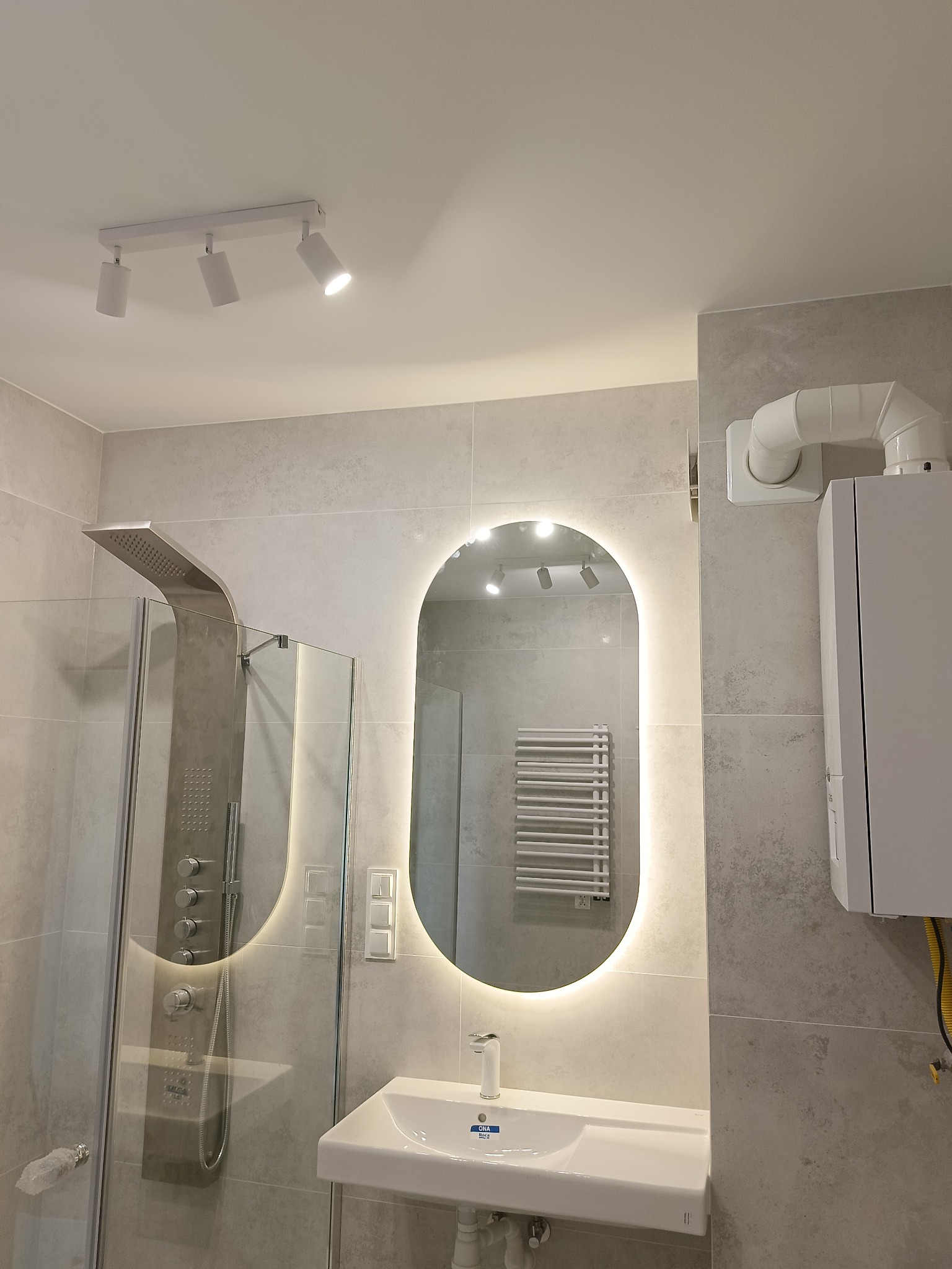 Nowoczesna łazienka z szarą glazurą, kabiną prysznicową z deszczownicą i podświetlanym lustrem owalnym nad białą umywalką. Widoczny bojler i oświetlenie sufitowe.