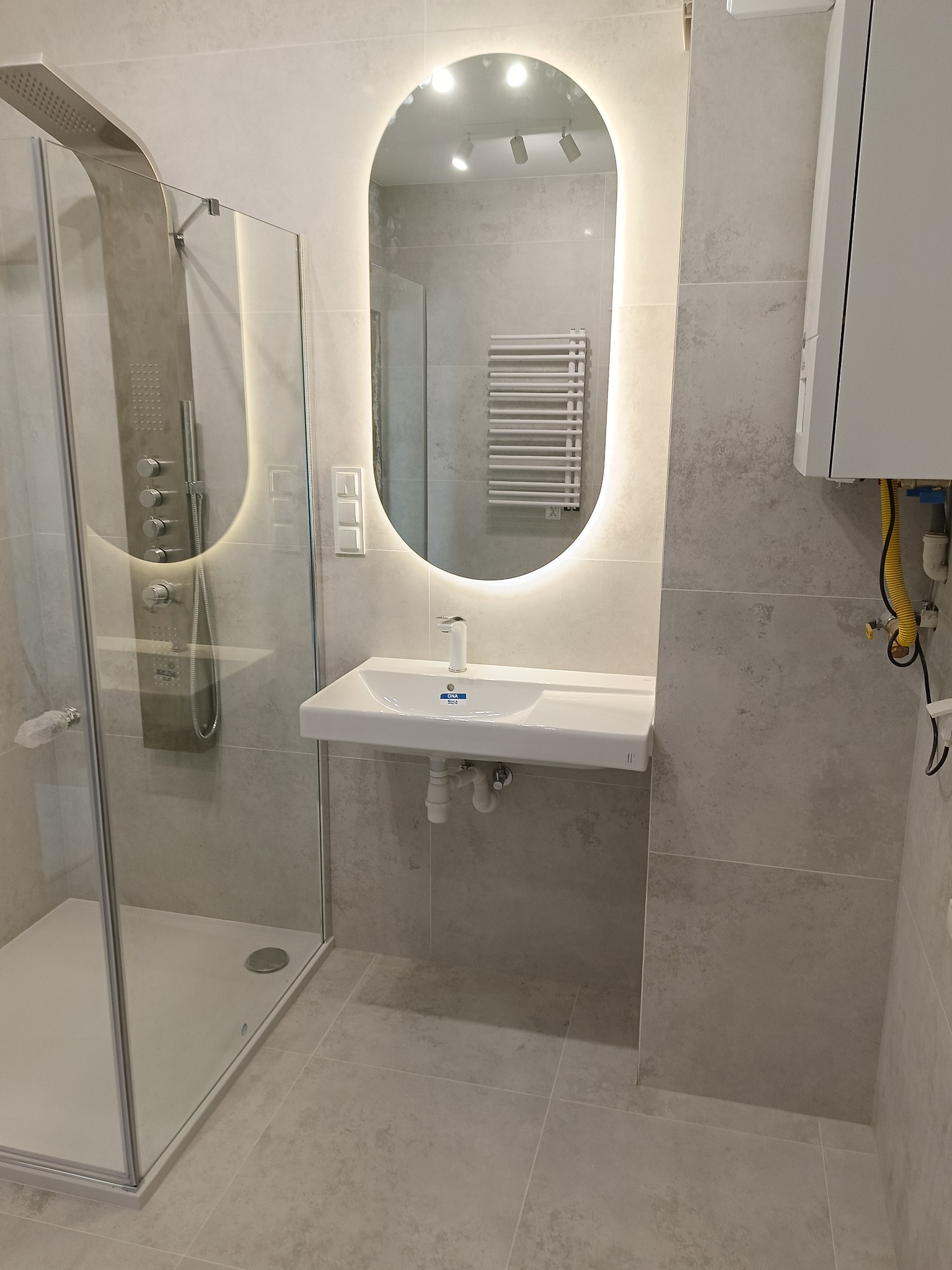 Nowoczesna łazienka w odcieniach szarości z kabiną prysznicową typu walk-in, podświetlanym lustrem i umywalką. Widoczny fragment pieca gazowego.