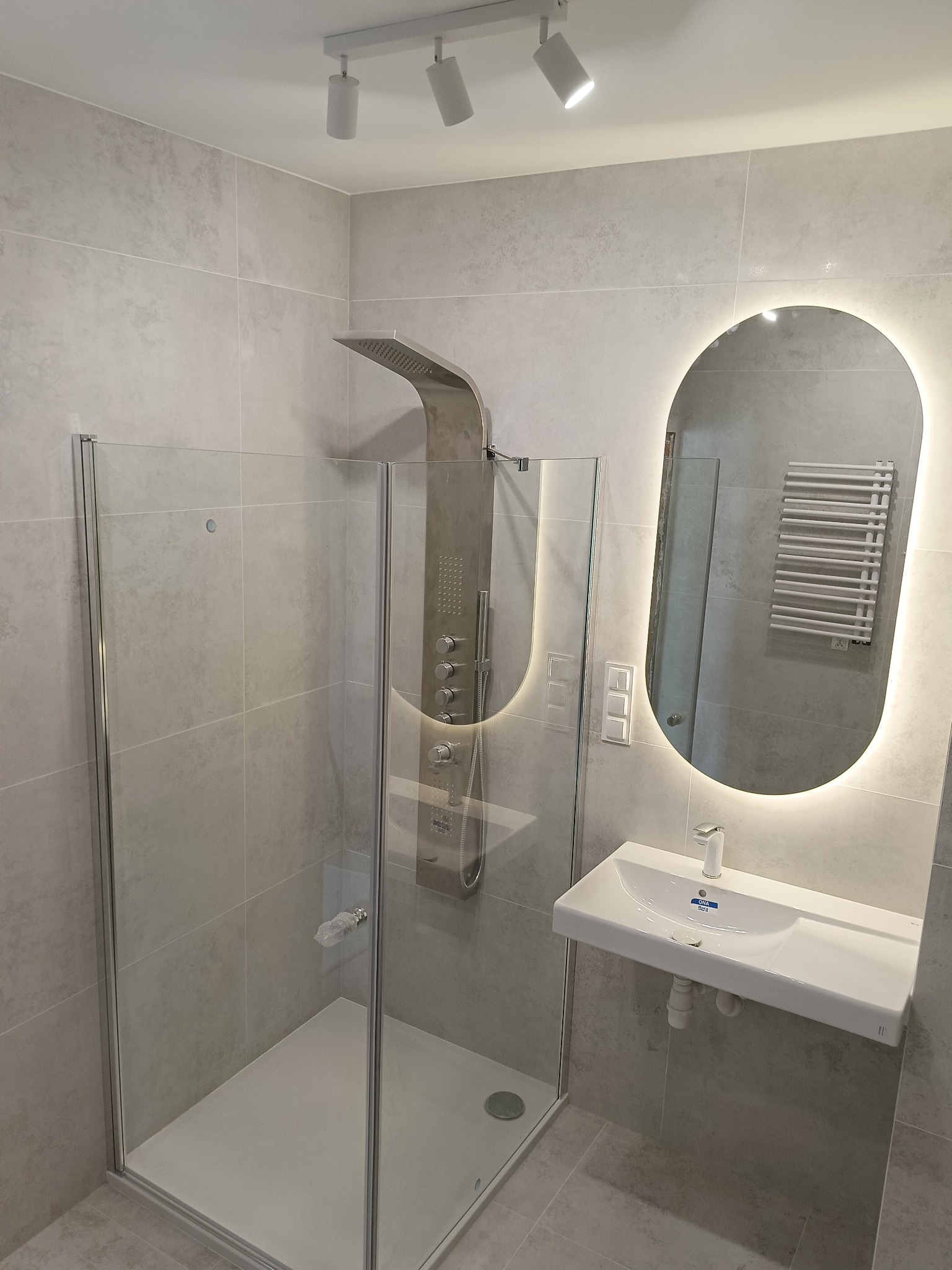 Nowoczesna łazienka z kabiną prysznicową typu walk-in, podświetlanym lustrem i umywalką. Szare płytki ceramiczne na ścianach i podłodze. Minimalistyczny design.