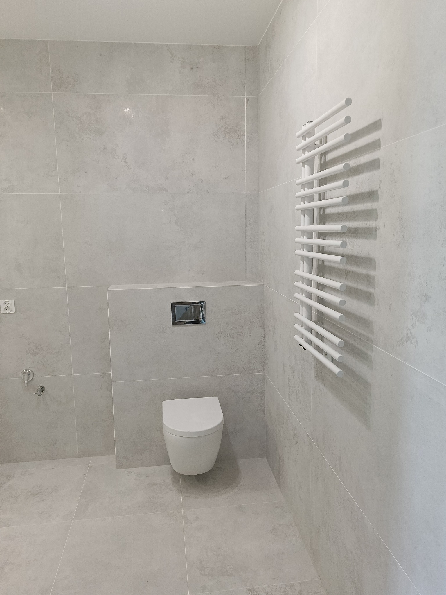 Nowoczesna łazienka w jasnych odcieniach szarości z wiszącą toaletą i białym grzejnikiem drabinkowym, wykończona płytkami imitującymi kamień.