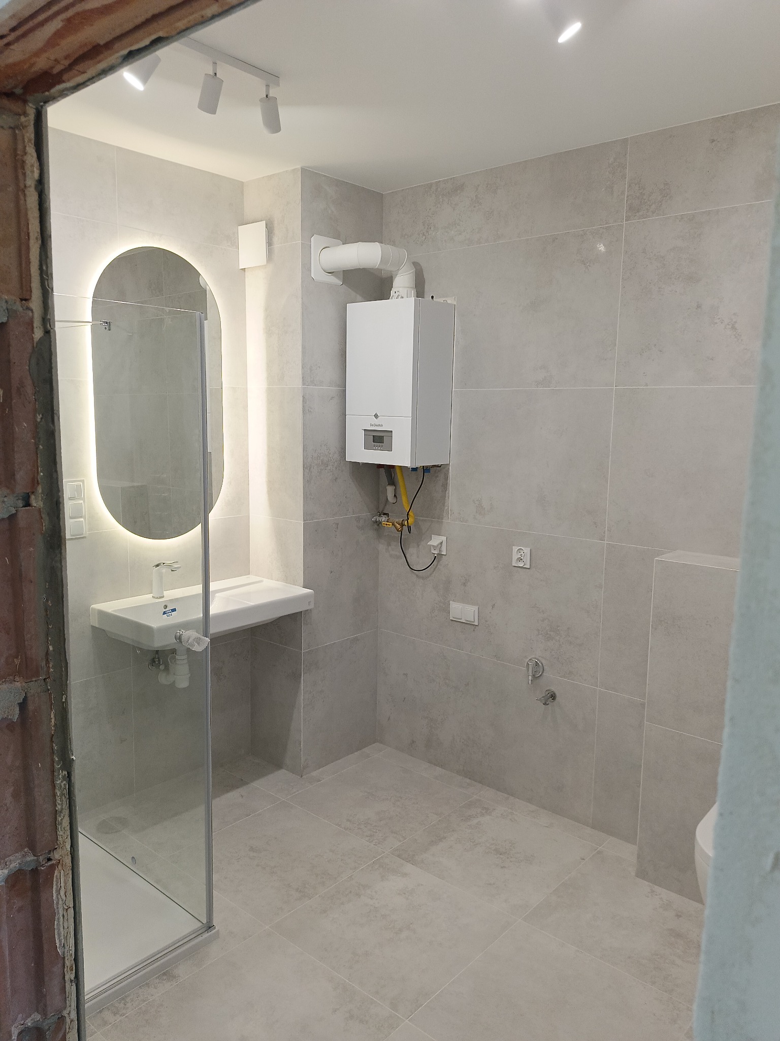 Wykończona łazienka w szarościach z podświetlanym lustrem, kabiną prysznicową i wiszącym piecem gazowym. Widoczne płytki imitujące kamień i nowoczesne oświetlenie.