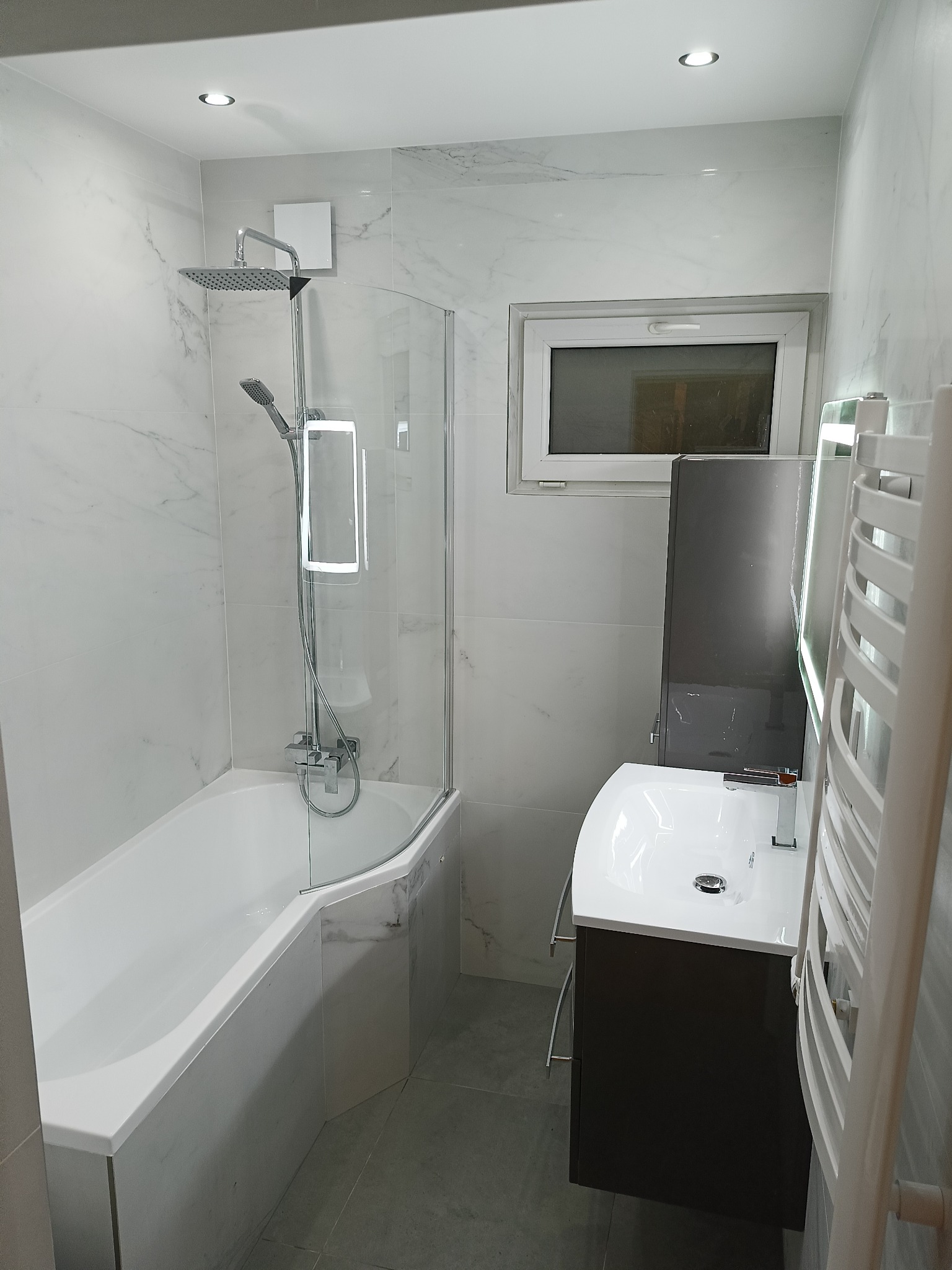 Nowoczesna łazienka z wanną z parawanem, umywalką z szafką i grzejnikiem drabinkowym. Białe marmurowe płytki na ścianach i szara podłoga.