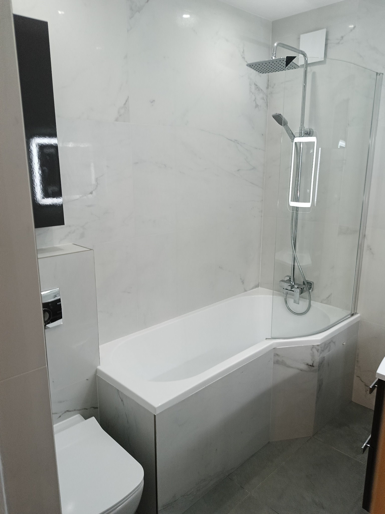 Biała łazienka z wanną obudowaną marmurowymi płytkami, szklanym parawanem i deszczownicą. Uwagę zwraca podświetlane lustro i szara podłoga.