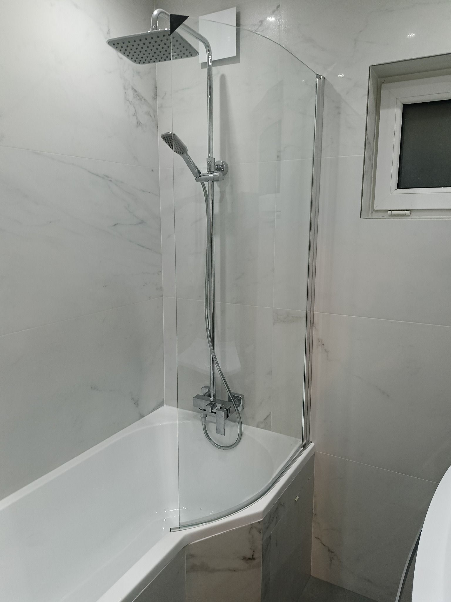 Biała wanna z zamontowaną szklaną ścianką prysznicową, wykończona marmurowymi płytkami. Widoczna bateria i słuchawka prysznicowa oraz okno w tle.