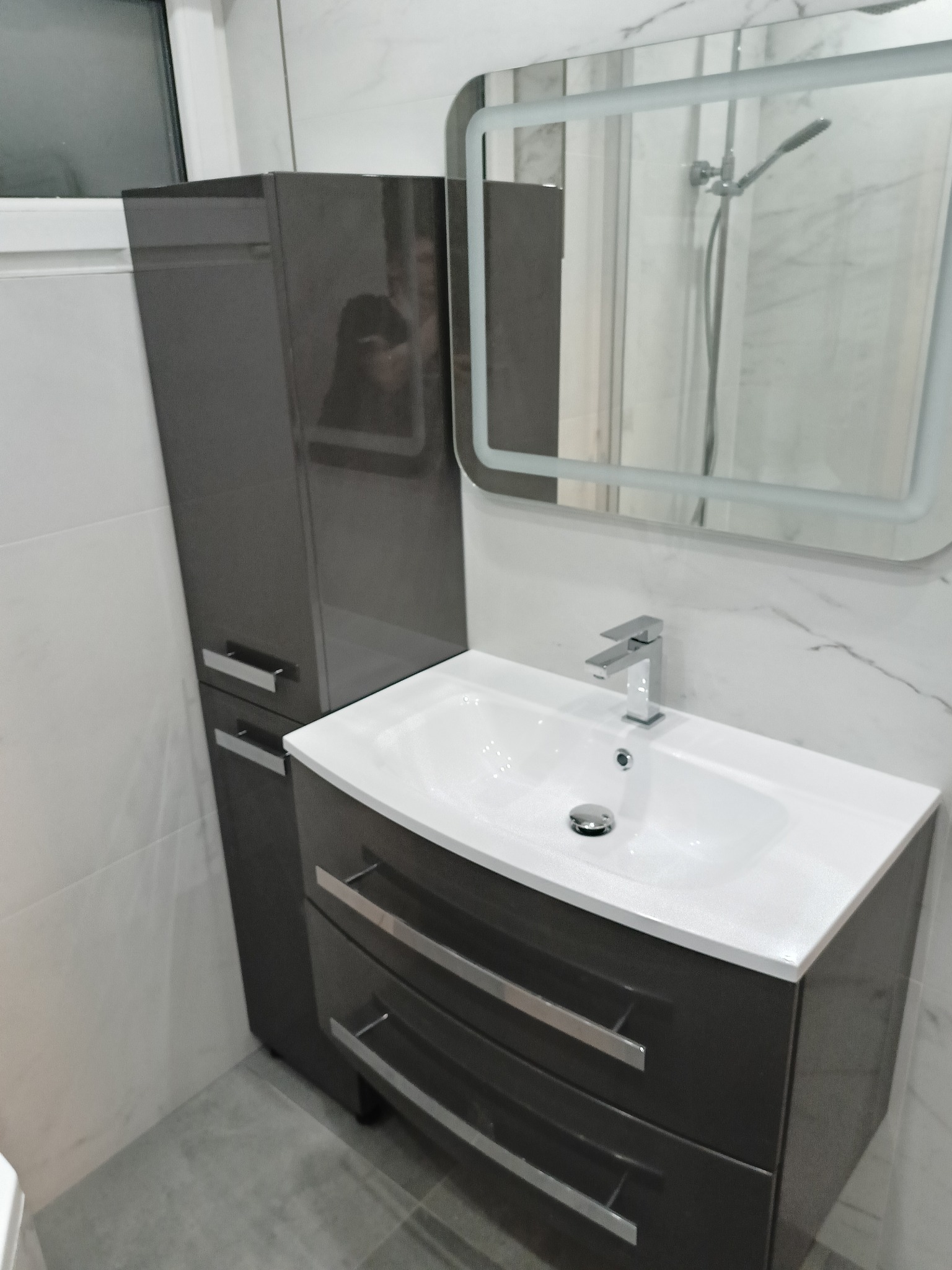 Nowoczesna łazienka z szarą szafką pod umywalkę, lustrem z podświetleniem LED i wysoką szafką. Białe płytki imitujące marmur na ścianach i szara podłoga.