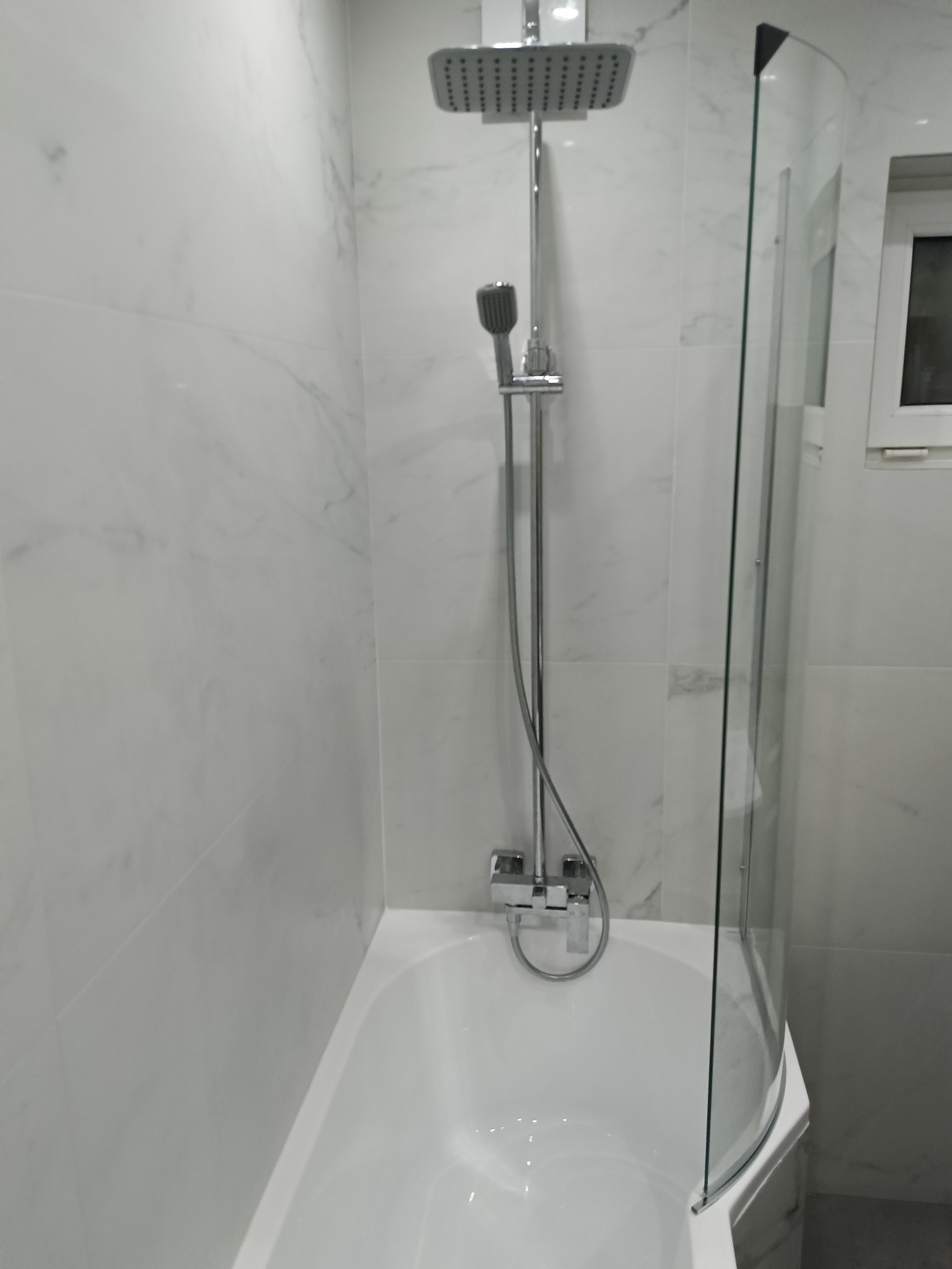 Biała, wykończona łazienka z wanną, szklanym parawanem i chromowaną armaturą. Ściany wyłożone płytkami imitującymi marmur. Widok na kabinę prysznicową.