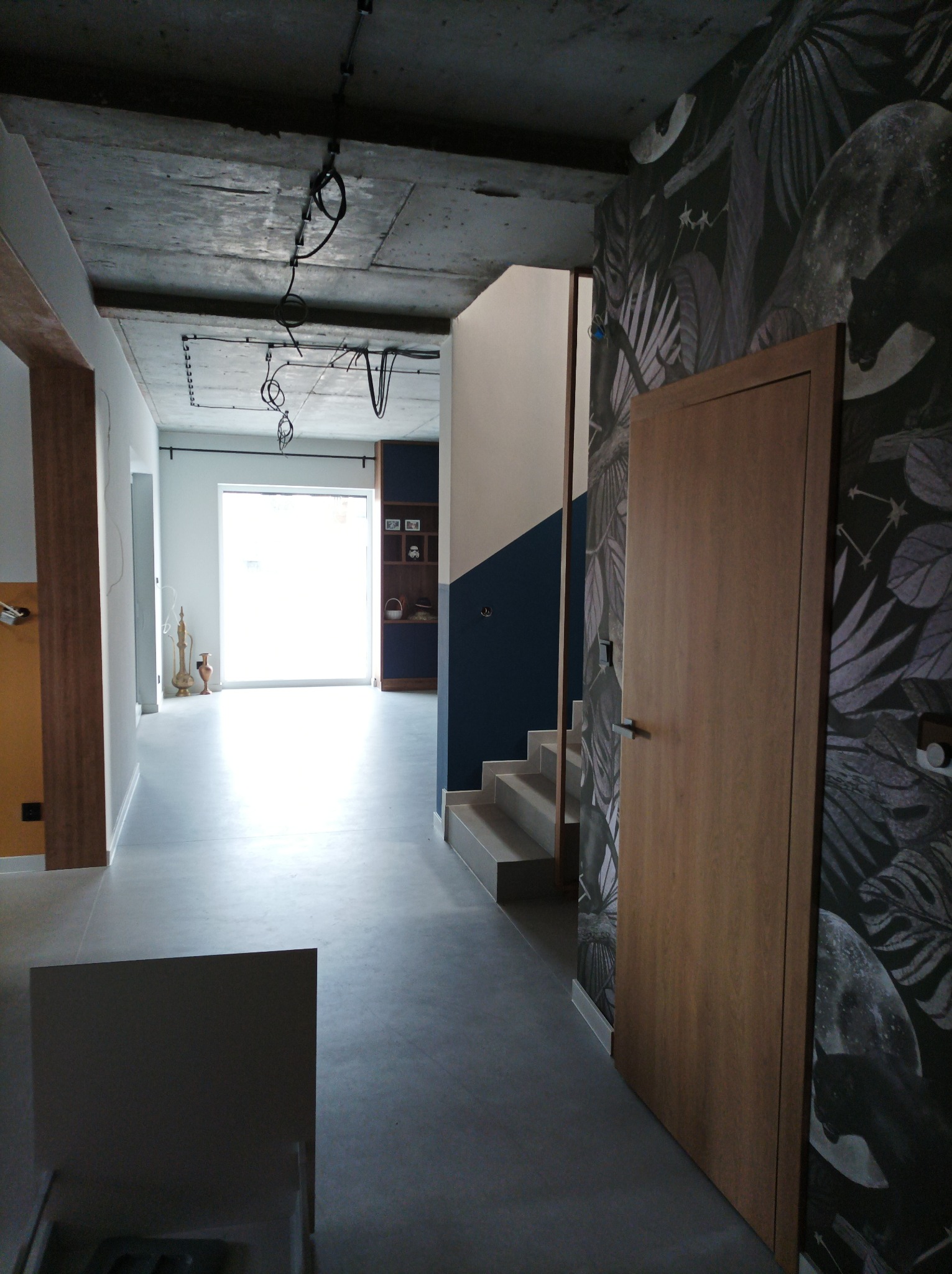 Nowoczesne wnętrze domu z surowym betonowym sufitem, szarą podłogą, schodami z granatowym akcentem na ścianie, drzwi z drewnianą okleiną i tapetą z motywem roślinnym