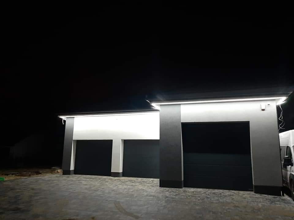 Nowoczesny budynek garażowy z dwoma bramami segmentowymi w kolorze antracytowym, oświetlony zewnętrznymi lampami LED pod okapem, sfotografowany nocą.