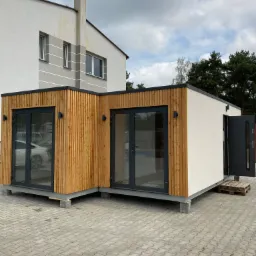 Nowoczesny pawilon z drewna i tynku z dwoma parami przeszklonych drzwi i otwartymi drzwiami wejściowymi, ustawiony na kostce brukowej przed budynkiem mieszkalnym.