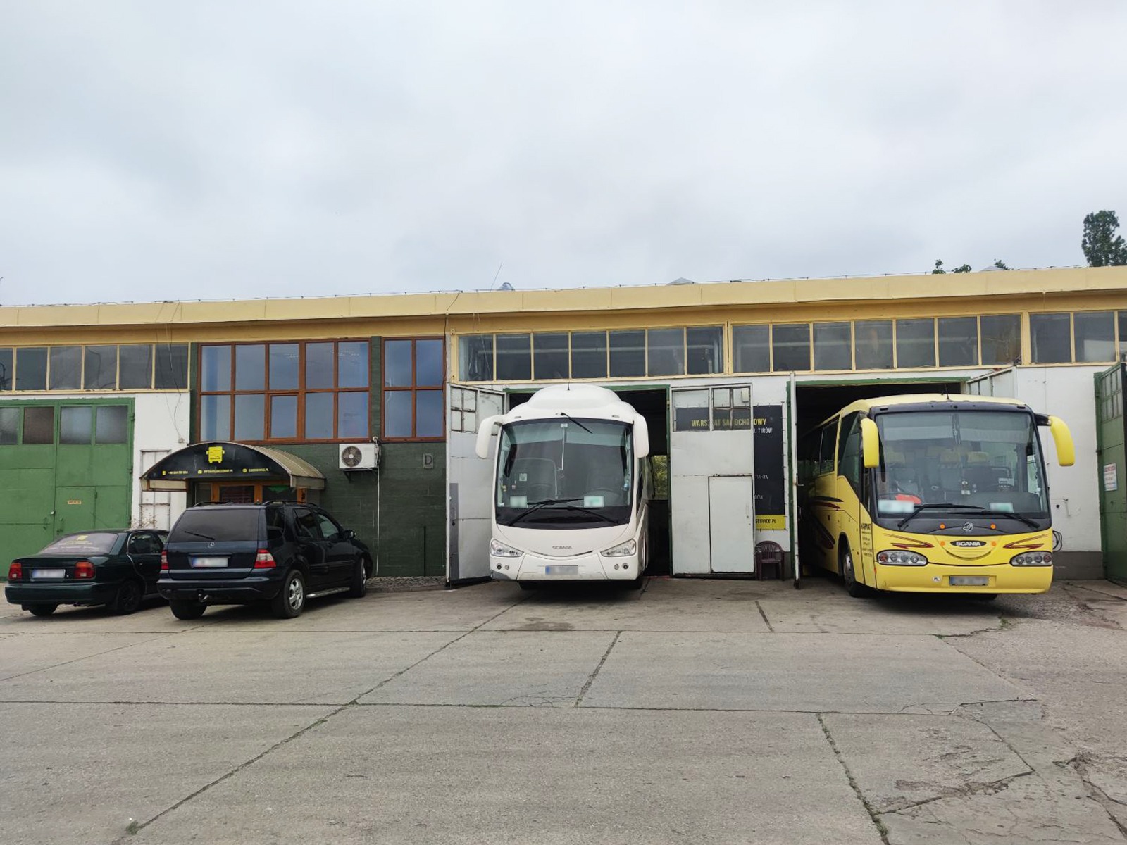 Dwa autobusy, biały i żółty, stoją w otwartych wrotach warsztatu samochodowego; obok zaparkowane dwa samochody osobowe; pochmurne niebo.