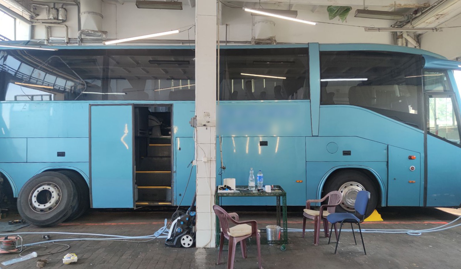 Niebieski autobus w warsztacie, z otwartymi drzwiami, widoczne opony, stolik z butelkami wody i krzesłami, narzędzia i kable leżące na podłodze.