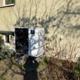 Biała, zewnętrzna jednostka pompy ciepła Mitsubishi Electric Zubadan R32, zainstalowana na betonowych podstawach przy ścianie budynku z widocznym cieniem drzewa.