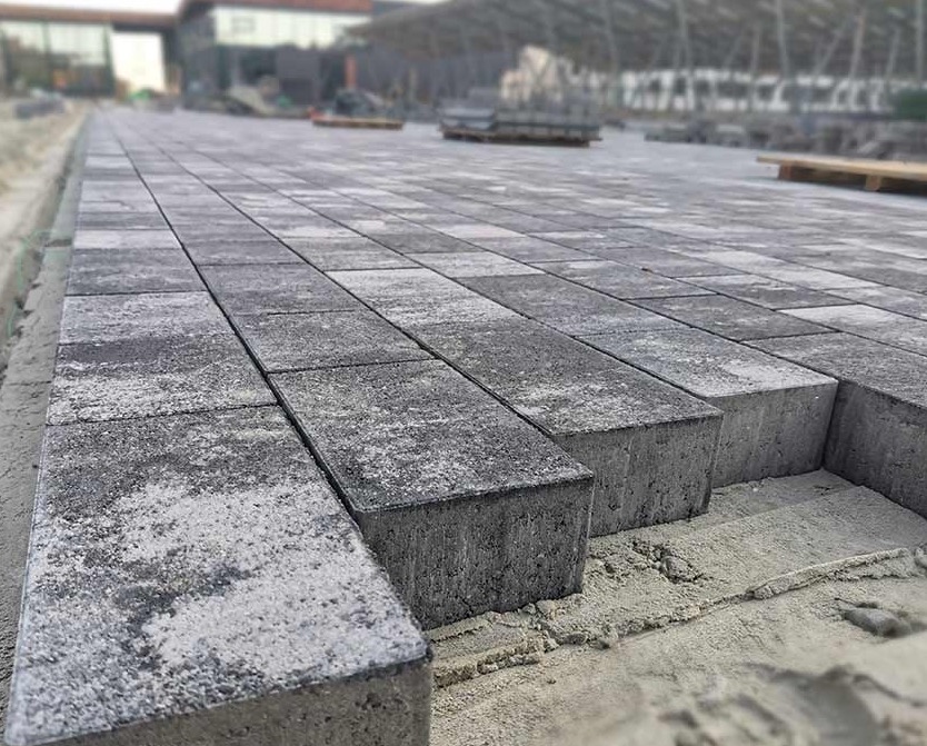 Układanie szarej, prostokątnej kostki brukowej na podsypce piaskowej, widoczne nierówności i niedokończony fragment nawierzchni.