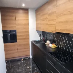 Nowoczesna kuchnia z czarnymi błyszczącymi płytkami podłogowymi imitującymi marmur i drewnianymi szafkami, czarny blat i czarne płytki trójkątne na ścianie nad blatem.