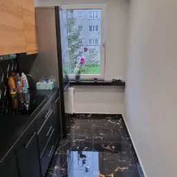 Czarne, marmurowe płytki podłogowe w kuchni, odbijające światło okna i lamp sufitowych, widok na zabudowę kuchenną z czarnymi frontami i drewnianymi szafkami wiszącymi.