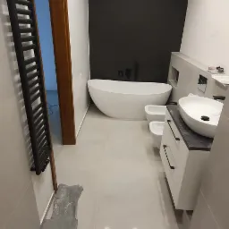 Nowoczesna łazienka z wolnostojącą wanną, bidetem, umywalką na szafce i grzejnikiem drabinkowym, wykończona w szarościach z czarnym akcentem ściany.