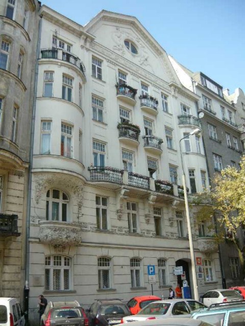 Elewacja kamienicy w stylu secesyjnym z balkonami i dekoracyjnymi detalami, widok z poziomu ulicy z zaparkowanymi samochodami.