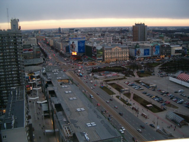 Panorama miasta z wysokości, widok na szeroką aleję z ruchem samochodowym, budynki biurowe i parking, oświetlone billboardy reklamowe, szara kolorystyka wieczornego nieba.