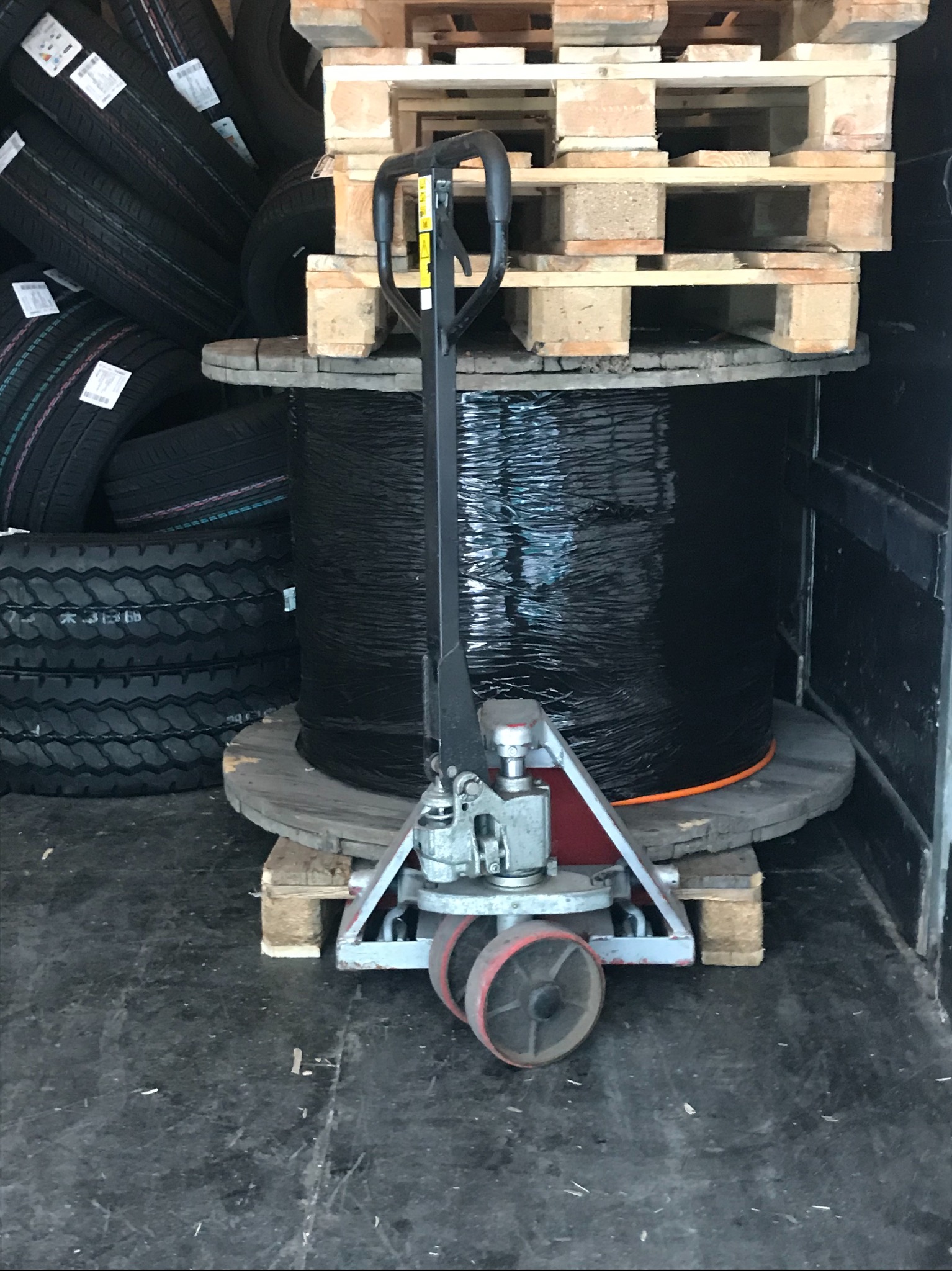 Wnętrze busa: palety, opony i wózek paletowy z dużą szpulą czarnego kabla na drewnianej podstawie, zabezpieczoną folią.