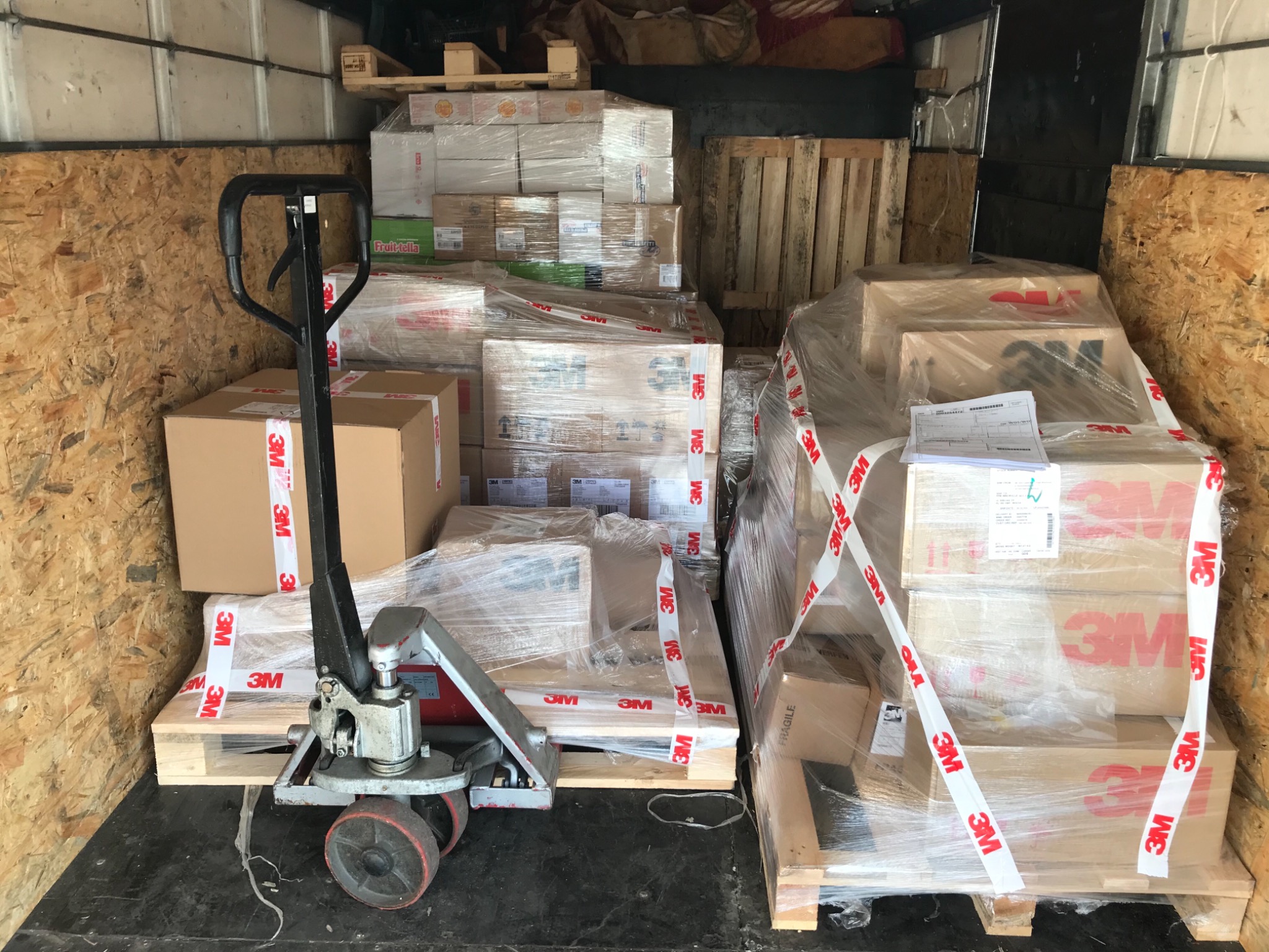 Wnętrze furgonetki załadowane paletami z kartonami zabezpieczonymi folią i taśmą 3M, z wózkiem paletowym z lewej strony, gotowe do transportu.