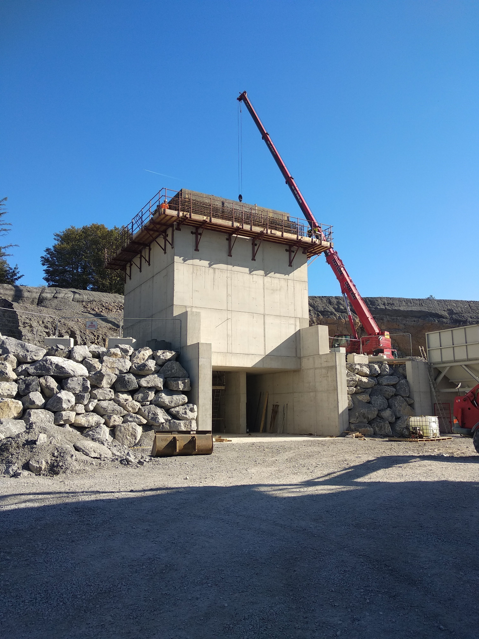 Wznoszenie betonowej konstrukcji z tunelem, widoczny dźwig teleskopowy i robotnicy na górnej platformie, teren budowy z kamiennym nasypem i sprzętem.