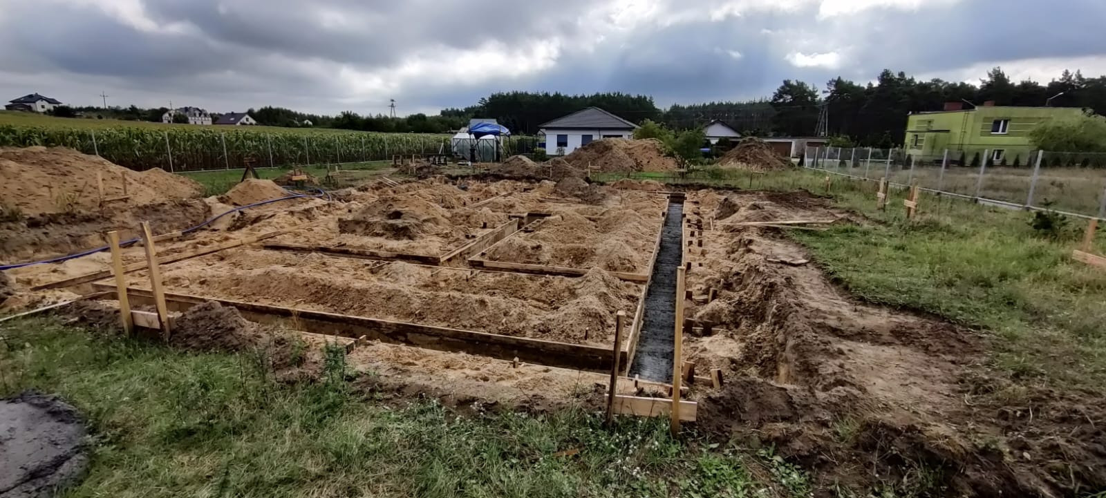 Przygotowanie fundamentów pod budowę domu jednorodzinnego w Bydgoszczy: wykopane rowy zalane betonem, szalunki z desek, widok na pole kukurydzy i domy w tle pod pochmurnym niebem.