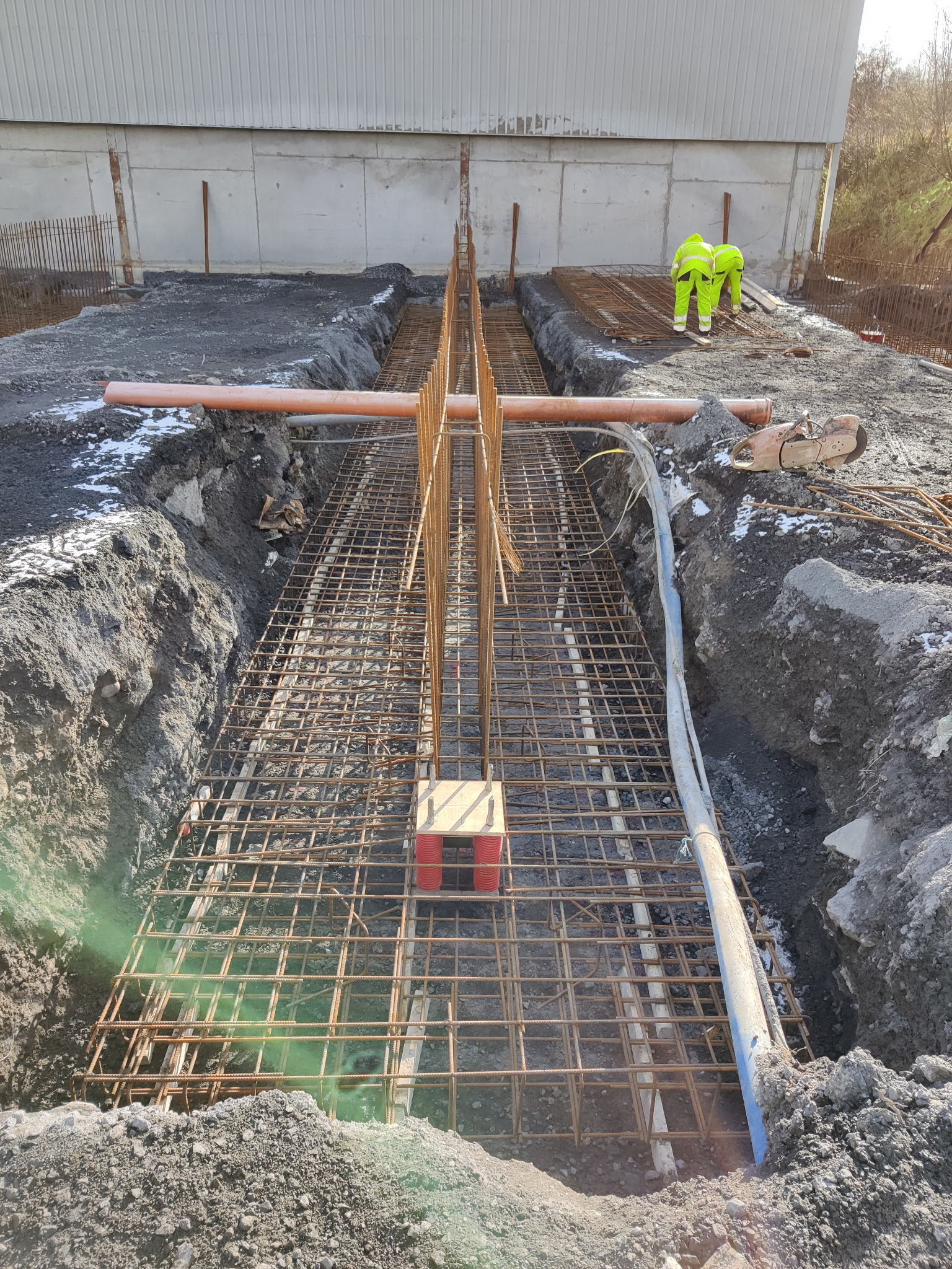 Przygotowanie zbrojenia fundamentów pod budowę hali, widoczne wykopy, siatka zbrojeniowa z prętów stalowych, rury oraz dwóch pracowników w odblaskowych ubraniach.