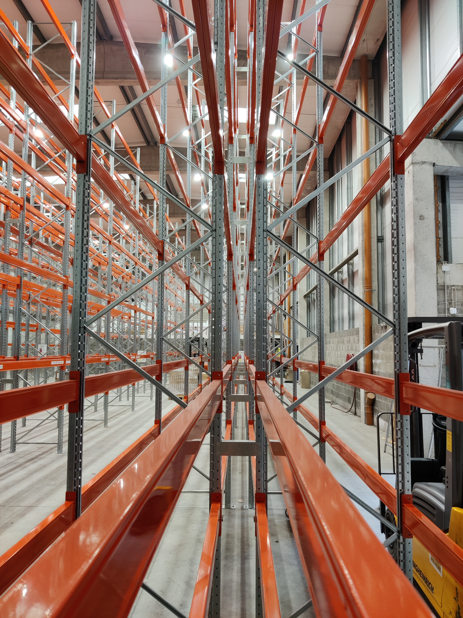 Wnętrze hali magazynowej z zamontowanymi, pomarańczowo-szarymi regałami wysokiego składowania, widok z perspektywy alei między regałami, z widocznym w tle wózkiem widłowym.