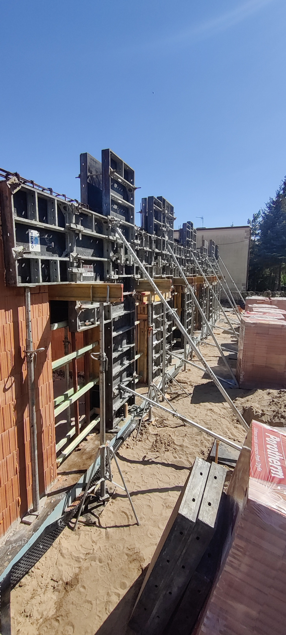 Budowa domu z czerwonej cegły Porotherm, widoczne szalunki i podpory stalowe na tle błękitnego nieba.