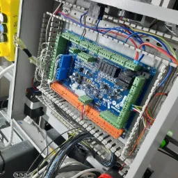 Otwarta skrzynka sterownicza z widoczną płytą główną, licznymi przewodami i złączkami, z żółtym przyciskiem awaryjnym po lewej stronie, oraz syreną alarmową na dole. Widoczne elementy elektroniki...