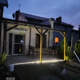 Oświetlony taras z drewnianą konstrukcją i meblami ogrodowymi, widoczny o zmierzchu, z dodatkowym oświetleniem LED pod dachem.