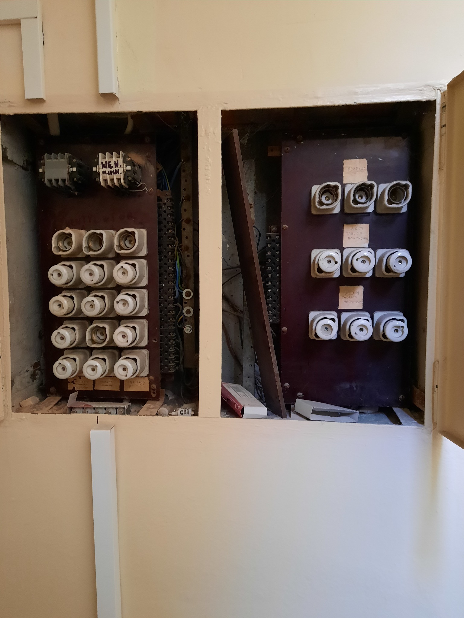Otwarta szafka z dwoma starymi tablicami bezpiecznikowymi z ceramicznymi wkładkami topikowymi, widoczne okablowanie i pozostałości po demontażu.